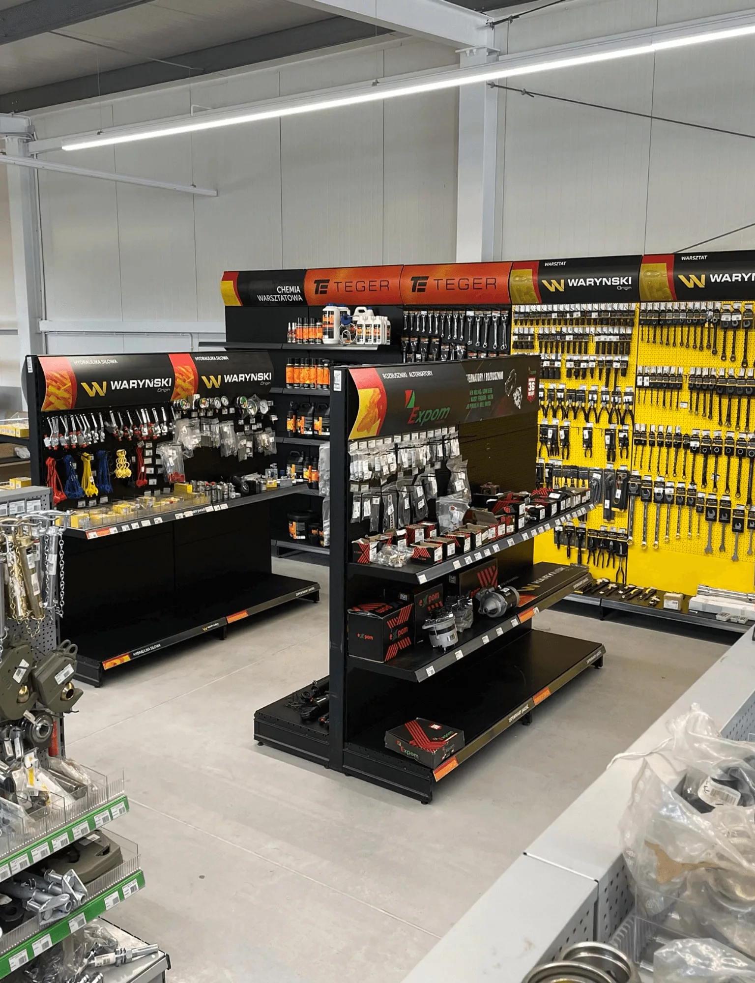 Warynski brand tools display in the shop, containing black-yellow shelves with organized tools on perforated wall. Yellow pry bars and loader ribs visible on the right, Warynski logo and branding above the shelves