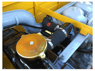 Close-up of a classic car's engine bay, showing a golden radiator cap, black rubber cooling hoses with a red logo, and a yellow reinforcement bar.